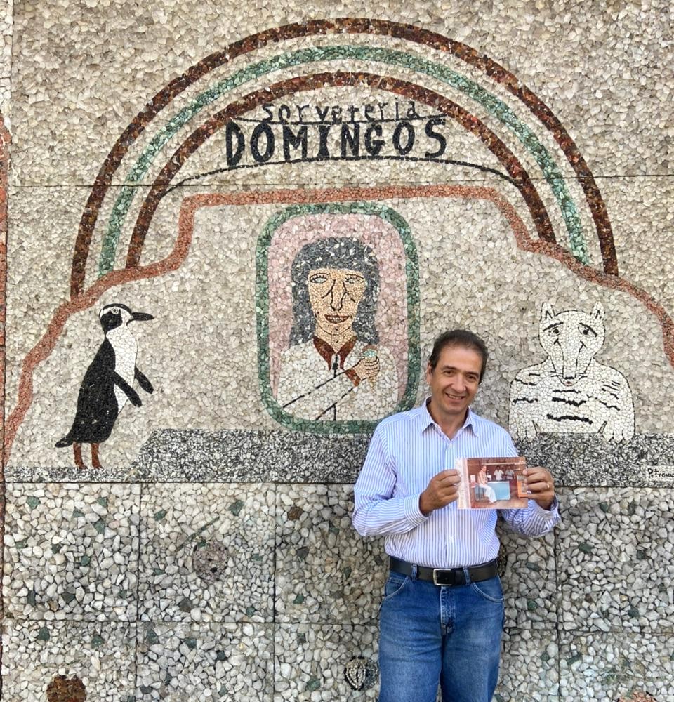 homem de pele branca, meia idade, segurando uma foto, em frente a uma sorveteria (fundo com parede cinza, com textura de pedra, alguns desenhos e texto: sorveteria domingos)